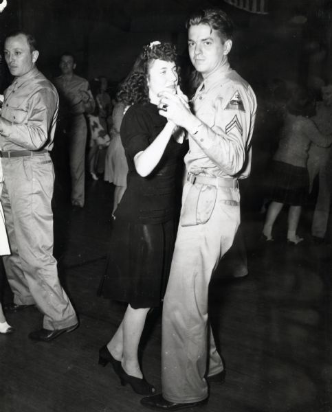 Uniformed Corp. J.L. Slack, a former International Harvester Company employee, dances with a woman wearing a dress and high heeled shoes at Port Clinton. Other dancing couples are in the background. Corporal Slack likely belonged to the "Harvester Battalion", and may have been stationed at Camp Perry, Ohio for training. The original caption reads: "LIGHT MOMENTS FOR A CORPORAL. Corp. J.L. Slack, formerly of the Chicago (m.t.) branch, joins the throng at the Friday-night dance conducted by the Port Clinton Service Center."