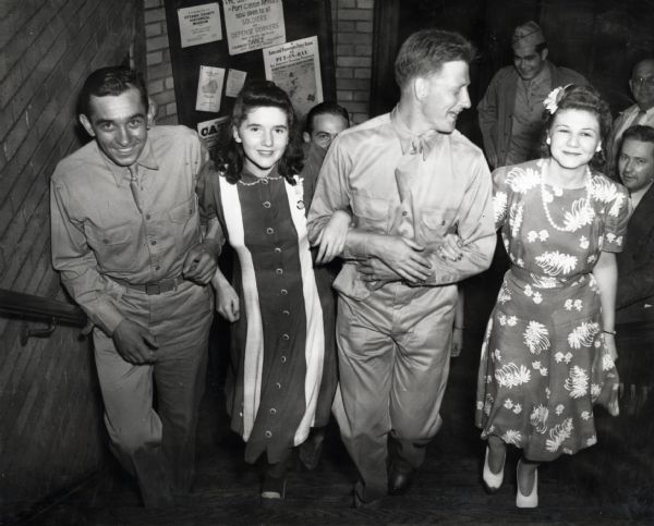 Two soldiers link arms with two women wearing dresses at the Port Clinton Service Center. The men were former International Harvester employees, and likely belonged to the "Harvester Battalion." The men may have been stationed at Camp Perry, Ohio for training. The original caption reads: "PLENTY OF FUN HERE. Coming up the steps at Port Clinton Service Center are (left to right): Corp. Joe E. Grewick, formerly of McCormick Twine Mill, Chicago: Katie Walters, Port Clinton, Ohio; Sgt. Wm. C. Newman, Minneapolis branch; Kay Zipperly, Cleveland, Ohio."