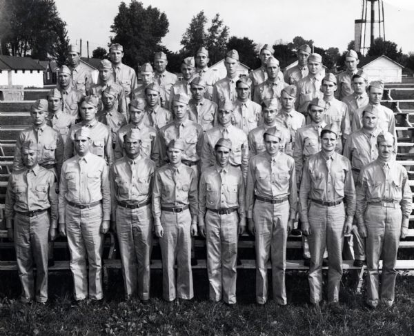 A group of uniformed enlisted men affiliated with International Harvester stand on risers for a group portrait. The original caption reads: "BATTALION FROM 40 STATES. Significant of the nation-wide response on the part of Harvester men to the call for formation of the "Harvester Battalion" is the fact that battalion members are from 40 states. The battalion men in the above group were selected to represent their home states in the picture."  The caption also lists the names of the men pictured. The photo may have been taken at Camp Perry, Ohio.