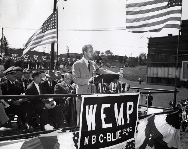 Fowler McCormick, president of the International Harvester Company, stands at a podium to address the audience at the Milwaukee Works "E" award ceremony. The ceremony is likely taking place near the factory. A sign advertises the radio station WEMP.