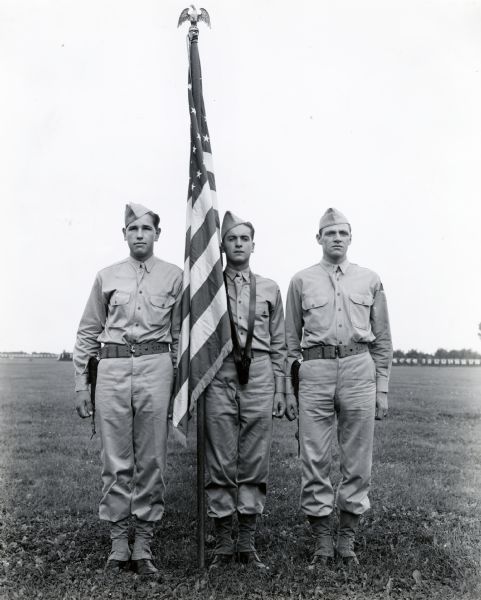 Three soldiers stand with an American flag in a field at Camp Perry. The men may have belonged to the "Harvester Battalion."