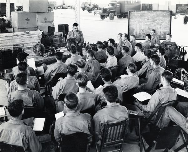 A group of soldiers gather in a large classroom at Camp Perry for a class on crawler tractors. A blackboard and various military vehicles are in the background. The men likely belonged to the "Harvester Battalion," which was made up of International Harvester employees and dealers.
