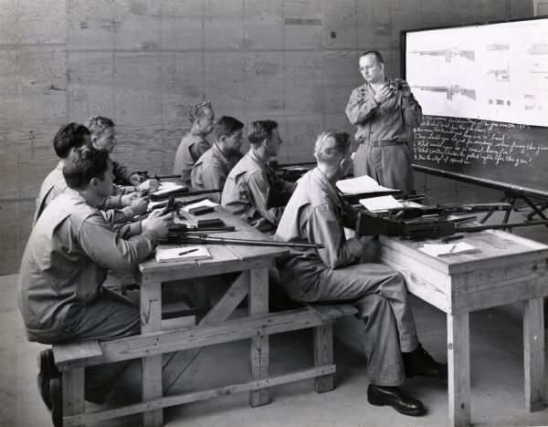 A group of uniformed soldiers sit at tables in a room at Camp Perry to be instructed in a small arms class. Firearms are on the table in front of each person and a chalkboard with script and illustrations on it is in the background. The soldiers likely belonged to the "Harvester Battalion," which was made up of International Harvester employees and dealers.