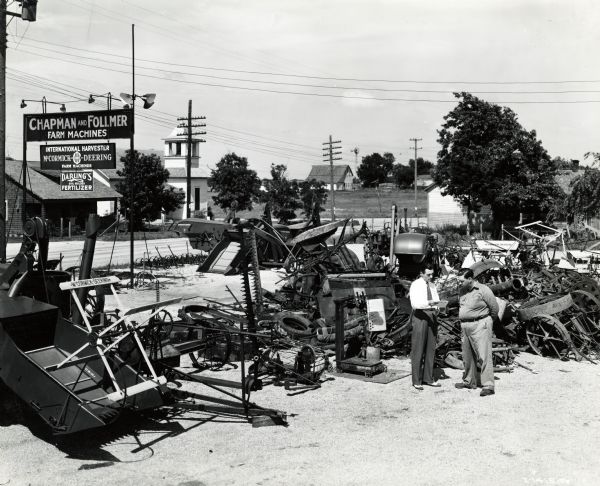 Two men stand outdoors by a pile of scrap metal intended for donation to the war effort at the Chapman and Follmer International Harvester dealership. A road, church, and farm buildings are in the background. The original caption reads: "Memory Jogger - Chapman and Follmer (E.F. Follmer, right), Avilla, Missouri, Springfield, Missouri, territory, pending disposition of this great pile of scrap, is using it as a daily reminder to all passers-by that scrap still at home can do the war effort no good until it gets to market and to the steel mills.  Assistant Manager L.J. De Mars, Springfield, Missouri, looks over the tonnage record of the firm and finds it good."