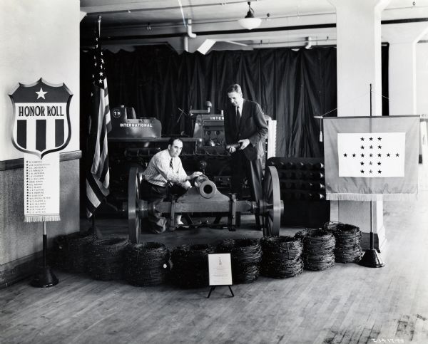 Two men assemble a patriotic display consisting of International machinery, a T-9 Tractractor (crawler tractor), a cannon, coils of barbed wire, signs, and flags at the Memphis branch of the International Harvester Company. The original caption reads: "Patriotic exhibit made of IHC parts at Memphis branch. I-1417-FF shows Floyd Sherrod (right), branch manager, and W.N. Felker, assistant manager."