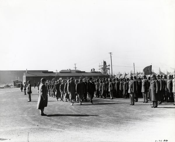 A group of soldiers stand at attention while lines of men walk past. Men are working on a roof and scaffold around a steeple on a building in the background. Rows of tents are in the background on the right.
