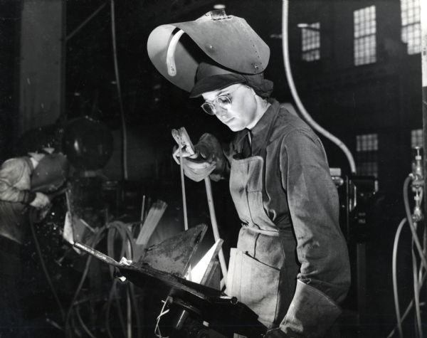 A female operator at an International Harvester Company factory welds the end plates of a M-5 high-speed tractor. The tractor was built for the Army to pull the guns of the Field Artillery. Another person is welding in the background.