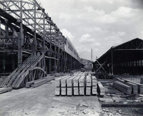 Stacks of lumber used in the construction of what is probably International Harvester's Bettendorf Tank Arsenal (factory). The lumber is sitting on the ground in front of incomplete building frames. A smokestack with vertical lettering reading "Bettendorf" is in the background. Men can be seen working around the building site.
