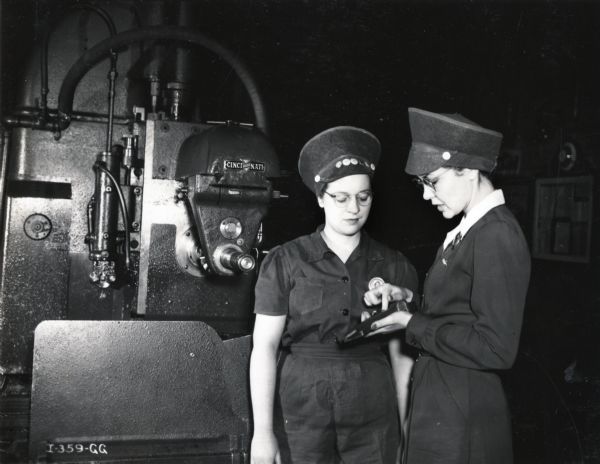 Two female factory workers talk while standing in front of a piece of machinery at International Harvester's Tractor Works factory. The original caption reads: "Gene Gerstynski, operator of a Cincinnati Milling Machine working on war production, confers with Genevieve Trumble, right, one of the women supervisors at International Harvester's Tractor Works in Chicago."