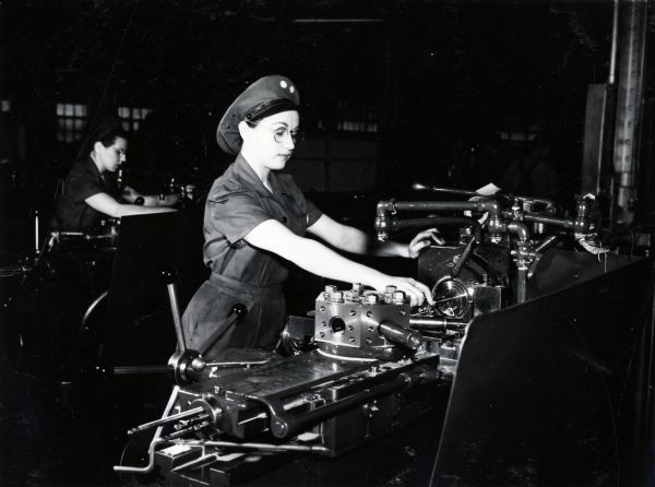 A female factory worker operates a lathe at International Harvester's Tractor Works. Another woman is working in the background. The original caption reads: "Women Employees in Harvester War Plants; Ernestine des Jardin, having completed her courses of training, operates a Warner-Swazey turret lathe used on war production at International Harvester's Tractor Works in Chicago."
