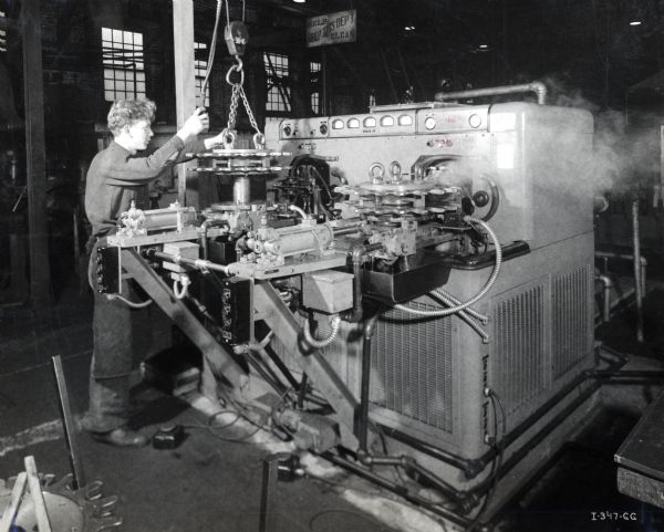 A factory worker using a Tocco induction process machine at International Harvester Company's Tractor Works. The original caption reads: "Operator readies one station on the Tocco induction process machine while the other station hardens a half-track sprocket. The Tocco process has increased the speed of this operation 2-1/2 times."