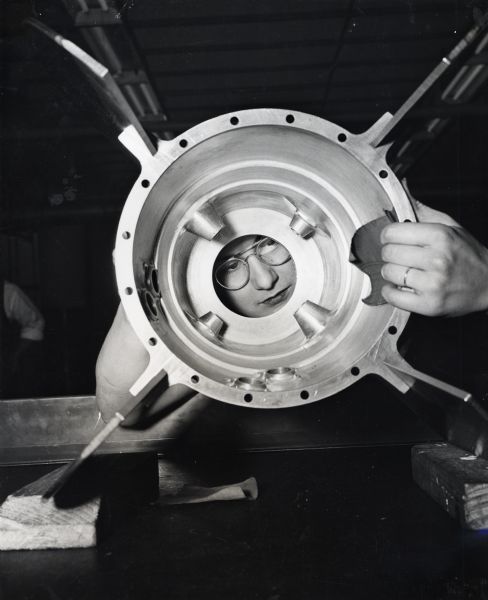 A female factory worker wearing safety glasses uses what appears to be a piece of sandpaper to polish a torpedo part at International Harvester's McCormick Works. Her face is visible through the hole in the torpedo. The original caption reads, "This woman employee at McCormick Works apparently has chosen the hard way of polishing a tail cone assembly of aircraft torpedo."
