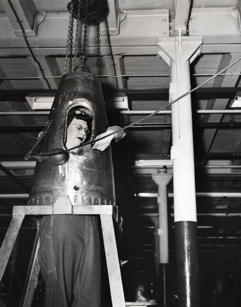 A woman's face is visible through an opening in the after-body of an aircraft torpedo as she uses a tool to tap holes in it. The photograph was taken at International Harvester's McCormick Works factory.