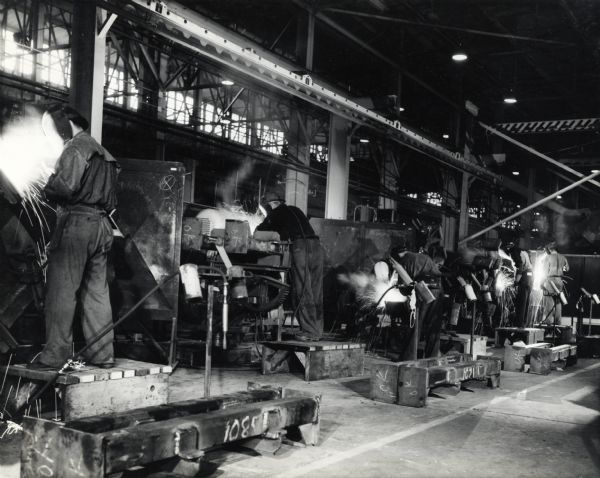 A line of factory workers at International Harvester's Bettendorf Works stand on wooden platforms at individual stations to weld parts for the "M-5 high speed tractor" [crawler]. The original caption reads: "These operators are welding the cross frame members of the M-5 high speed tractor at Harvester's Bettendorf (Iowa) Works. The fixtures permit tilting the members to the most advantageous position for each welding operation."
