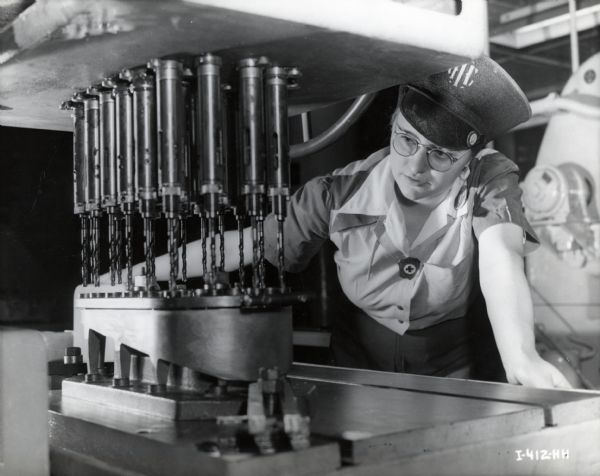 Factory worker Doris Zwieg uses a multiple drill for war production at International Harvester Company's Milwaukee Works. She wears a hat embroidered with the initials "IHC." The original caption reads: "Doris Zwieg carefully operates the multiple drill which bores holes in the base of a Gyro mechanism."
