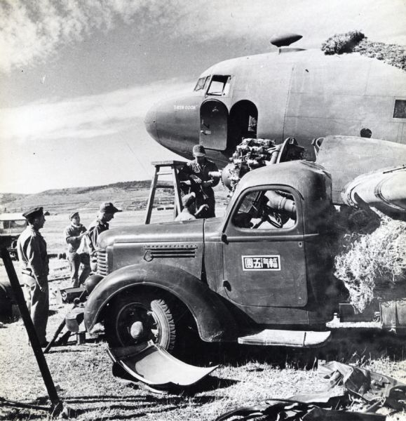 Uniformed soldiers use a ladder to unload objects from a U.S. Army C-47  airplane labeled "Green Goon." An International truck appears in the foreground with a panel of characters on the door.