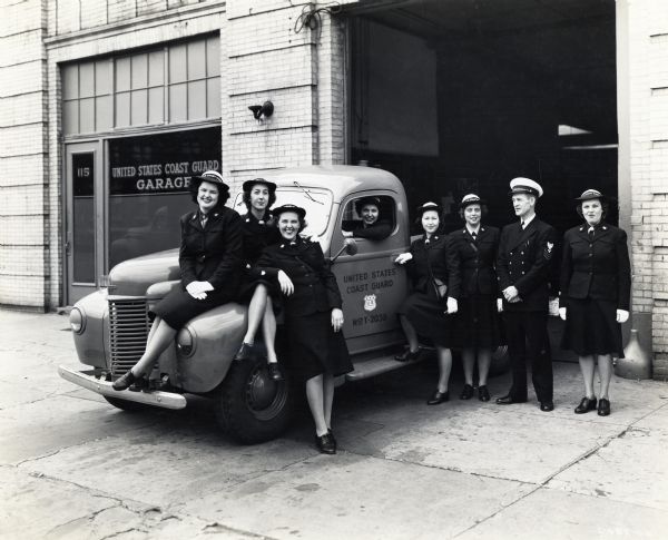 A group of uniformed women and a uniformed man stand around and sit on an International truck marked "United States Coast Guard" at the entrance to the United States Coast Guard garage.