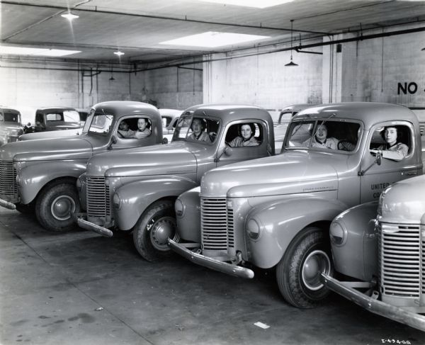 Six women sit in the driver and passenger seats of three International trucks used by the United States Coast Guard. The trucks are parked indoors in what appears to be a parking structure, possibly at a United States Coast Guard garage.