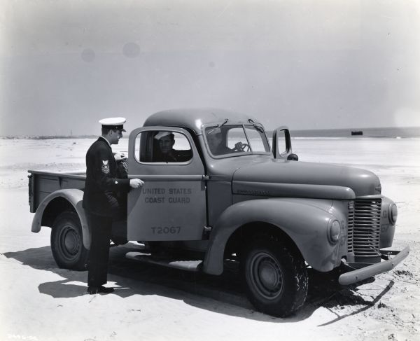 Two uniformed men sit inside an International K-1 U.S. Coast Guard truck parked on a beach while another man speaks to them through the open passenger door. The original caption reads: "Oversize Tires for Trucks on Beach Patrol: To the fighting United States Coast Guard is assigned the sizable task of guarding our far-flung shore lines. Close watch is continuously maintained from out-of-way watch towers and by armed beach patrols. Vehicles which serve the numerous beach outposts are equipped with oversize tires as shown on the half-ton International pickup in the illustration to facilitate movement in sand."