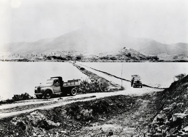 An International K-5 truck and an M-2-4 truck used by the Seabees are driving along a dirt road built over a body of water.