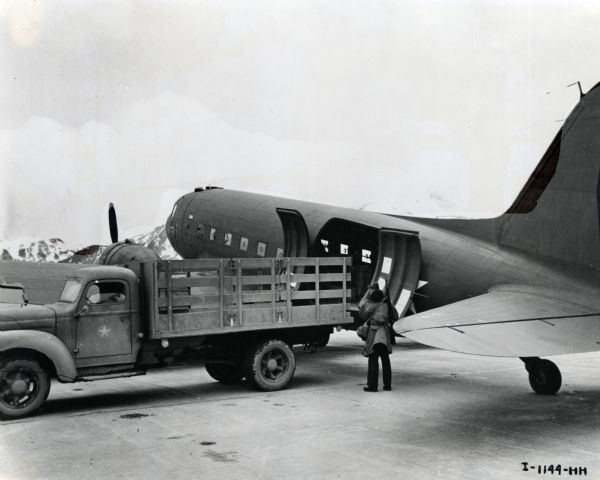 A man supervises the unloading of a US Navy R4D transport plane onto a truck with a star symbol painted on the driver door. Snow-capped mountains are in the background. The original caption reads: "TRANS-SHIPPING---Under the watchful eye of a Marine sentry, a Navy transport plane is unloaded at an Aleutian airfield."