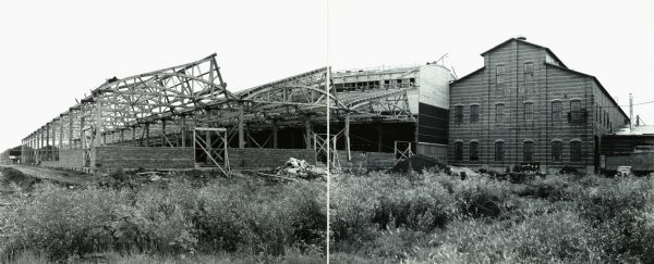Panoramic view of what appears to be an incomplete factory building beside a completed building marked "1910". A car is parked in the grass on the right. The buildings may be International Harvester's tank arsenal at Bettendorf, Iowa. The original caption reads: "Approved on 10/27/42 for use by McQuinn in Harvester News-Letter by G.B. Nidal, Chi. Ord. Dept."