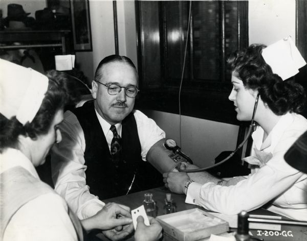 Two nurses test C.E. Stevens' blood pressure and hemoglobin prior to his blood donation at the Red Cross blood bank. The original caption reads: "Blood pressure and hemoglobin tests for C.E. Stevens, former assistant manager of motor truck sales, now manager, War Materials Division, International Harvester Company, prior to his blood donation at Red Cross blood bank, Chicago."