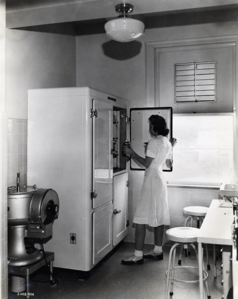 A woman in a white dress, probably a nurse or volunteer, places a container into a "Tomac Plasma Bank," a refrigeration unit developed by International Harvester.