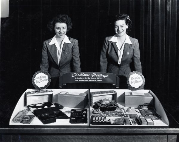Two women stand behind a display showing International Harvester's Christmas gifts for it's male and female employees in the armed forces. The women's gift includes a bag of candy or nuts, fudge, a toiletry set, and a sewing kit. The men's gift includes a checkers set, toiletries, a duffel bag, fudge, candy or nuts, and a sewing kit.