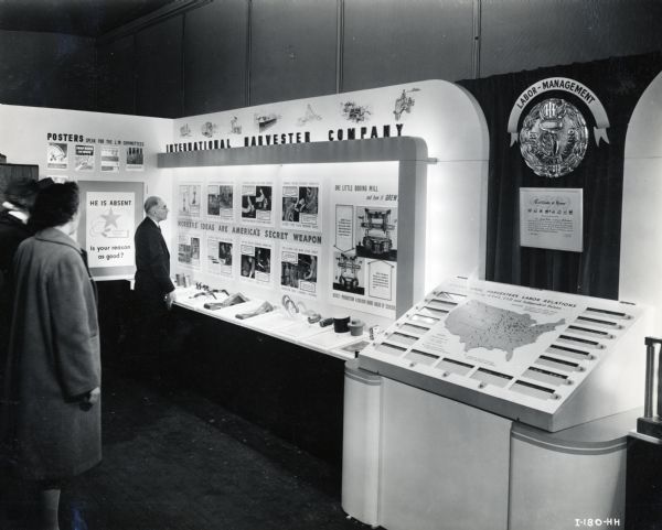 Men and women look at an International Harvester Company display featuring illustrations, posters, and machinery parts used in wartime work. The text in the display reads: "Workers' Ideas are America's Secret Weapon."