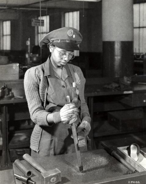 A woman wearing a hat, protective gloves, and safety glasses uses a tool to bend copper tubing used in making aircraft torpedoes at an International Harvester Company factory. The original caption reads: "International Harvester developed an important time-saving technique for the bending of copper tubing used in the torpedo. It is one of the most important contributions the Company made to the mass production of torpedoes. Large tubes, which are to be cold bent, are filled with silica sand before bending. Plugs are inserted in each end of the tube to keep the sand in place. This photograph shows a tube after it has been filled with sand and with the ends plugged."