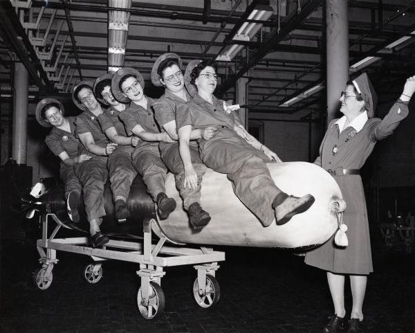 Side view of six women sitting on a torpedo placed on a rolling cart at an International Harvester factory. Another woman woman stands to the side with her left arm raised; they wear uniforms and hats embroidered with the IHC logo.