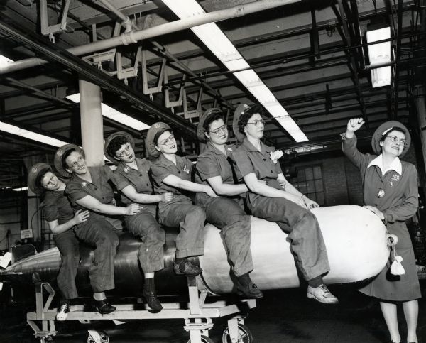 Side view of six women sitting on a torpedo placed on a rolling cart at an International Harvester factory. Another woman woman stands to the side with her right arm raised. They wear uniforms and hats embroidered with the IHC logo.