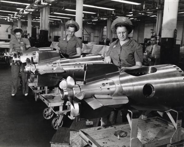 Three women work on a line of torpedoes at an International Harvester factory. The women wear uniforms and hats embroidered with the IHC logo. There is a man holding a camera is in the background.