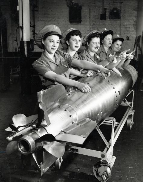 Five female factory workers stand beside a torpedo pointing their fingers and wearing IHC uniforms and hats to pose for a group portrait at one of International Harvester's factories.
