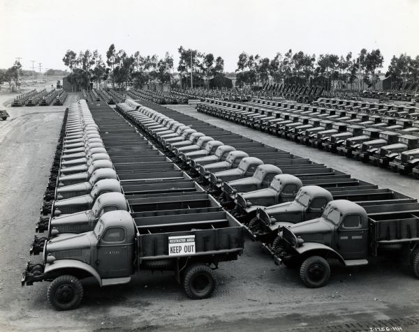Elevated view of International trucks parked in rows in a dirt lot after being prepared for overseas shipment to United States Seabees. The original caption reads: "After International trucks and tractors are given their various tests, and are sprayed with rust-preventing compound for oversea travel, they are lined up with military precision in solid ranks as shown and are ready for immediate embarkation and the numerous war zone activities in which the Seabees continually engage them. Here in the front two ranks, with thirty-one units in each rank are Model M-3H-4 four-wheel drive, cargo trucks, or as they say at the base four by fours. Immediately back of these are two more solid ranks of four by four M-3H-4 dump trucks. Back of these are more and more solid ranks of International trucks as far as the camera would let one see."