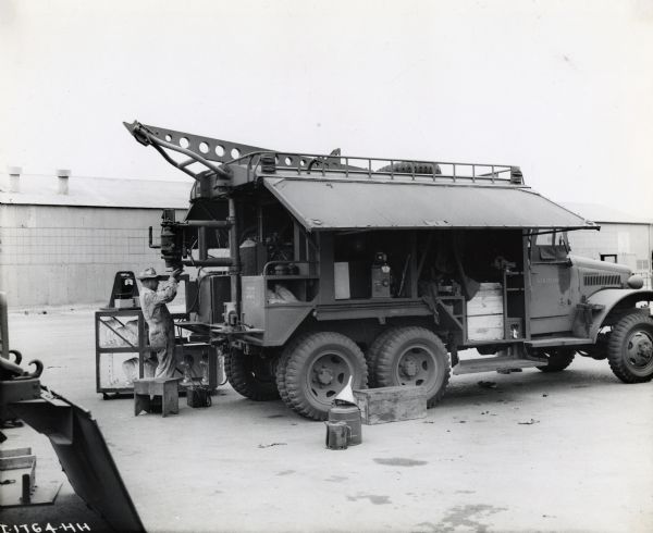 A man dressed in coveralls and a hat inspects a part on the rear of an International truck built for the U.S. Military. The side panel on the vehicle is lifted to reveal storage areas within the truck. There is a long building in the background. The original caption reads: "At the time, various pieces of apparatus were undergoing tests prior to shipment overseas. These mobile shops are designed especially for use in airfield work."