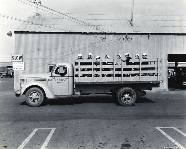 A group of uniformed men sits in the back of an International K-7 truck used by the U.S. Navy. The truck is driving in a parking lot in front of a large metal building. The original caption reads: "A number of standard K-7 Internationals with stake bodies are utilized at the Port of Hueneme for general hauling. Some of them are provided with seats as shown and serve as personnel carriers. At night these K-7's are parked in a group with other standard International models, as shown in photo I-1776-HH."
