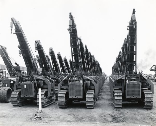 International crawler-tractors outfitted with Trackson cranes park in rows in a dirt lot. The original caption reads: "Just as is the case with International motor trucks described above, International crawler tractors receive careful tests and then are lined up in big groups according to models ready for shipment to the various Pacific battle areas."