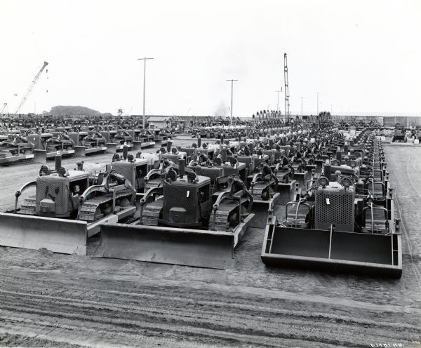 Elevated view of International TD-9 crawler tractors (TracTracTors) equipped with bulldozers and front-end loaders parked in rows in a dirt lot. The original caption reads: "Just as is the case with International motor trucks described above, International crawler tractors receive careful tests and then are lined up in big groups according to models ready for shipment to the various Pacific battle areas."