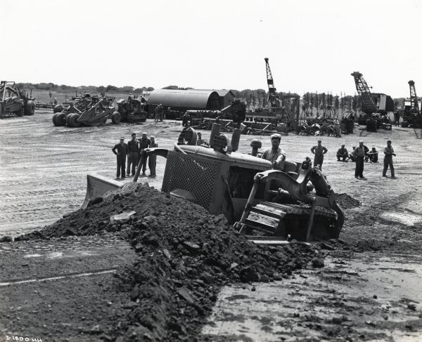 Seabees at Camp Rousseau train to use an International TD-18 diesel crawler tractor (TracTracTor) equipped with a bulldozer. Various vehicles and machinery are in the background. The original caption reads: "Part of the intensive training received by Seabees at Camp Rousseau, located in the Port of Hueneme area, is a thorough drilling in the operation of crawler-tractors. The illustrations show Seabees at work with International TD-18 Diesel crawler-tractor, equipped with bulldozer and demonstrating how it can be used to dig a trench, to build a mountain trail by angling the blade and to excavate burying places for fuel tanks. The pictures also show groups of white and colored Seabees receiving instruction on operation of the tractor."