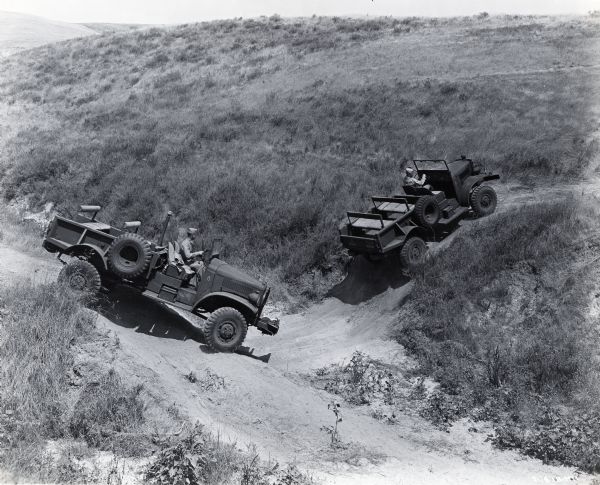 Two International trucks follow a dirt path into and out of a small valley at a Marine Corps Motor Transport School Training Command Camp. The school was located at Camp Joseph H. Pendleton. The original caption reads: "Most spectacular of the numerous uses to which International motor trucks are put at Camp Pendleton are those of four-wheel-drive and six-wheel-drive units in the Motor Transport School Training Command Camp. Some 45 trucks are continually employed in the training course and because of the unusually difficult operations which trainees must subject the trucks up and down near precipitous slopes, across deep gullies and in cross-country night driving without lights, wrecks are numerous and frequent replacements therefore are necessary. The training course lasts 8 weeks and training drivers grouped in two classes. The pictures both action and still (simulated action) show eight Model M-2H-4 four-wheel drive and two Model M-5N-6 six-wheel-drive Internationals with training Marine drivers at their wheels putting the trucks through their paces over very difficult mountain terrain."