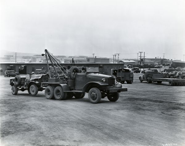 A man uses a truck marked "USMC 66750" to haul a wrecked vehicle through a parking lot at what might be Camp Pendleton. There are numerous vehicles and buildings in the background. The original caption reads: "Crack-ups of equipment caused by severe cross-country training practice require the use of a big six-wheel wrecker, a M-5H-5, which is shown pulling in a wrecked M-2H-5."