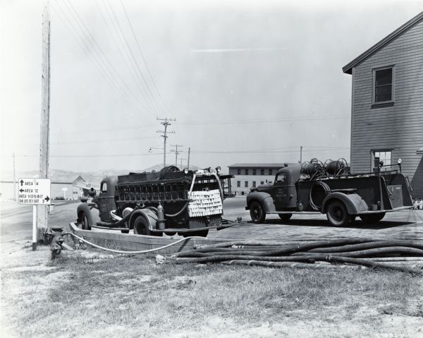 Two K-5 International trucks equipped with hoses and ladders used by the Camp Pendleton fire department are parked near a building and a road sign. The original caption reads: "A fire department is an important adjunct of Camp Pendleton. Here are shown two K-5 Internationals with hose and ladders, extinguishers and 350-gallon tanks with pressure pumps."