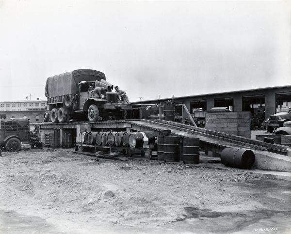 Two men performing service work on an International M-5H-6 six-wheel-drive truck with a cargo body parked on a grease rack outdoors. The truck is marked, "USMC 68436". Long buildings with open garage doors are in the background.