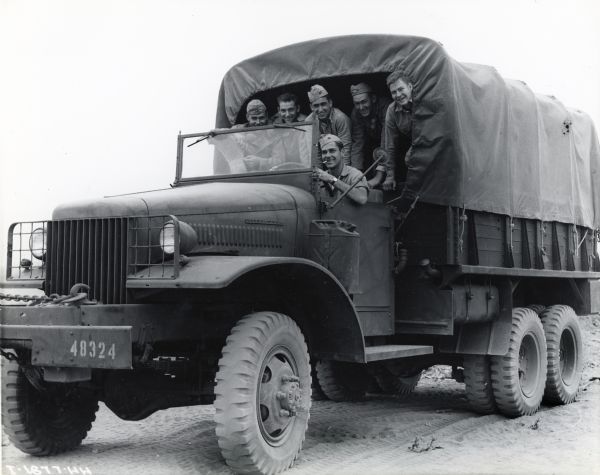 A group of five uniformed men stand in the covered cargo body of an International M-5H-6 six-by-six truck with a driver smoking a cigarette sitting behind the steering wheel. The original caption raeds, "M-5H-6 "six by six" International with cargo body and trash gang."
