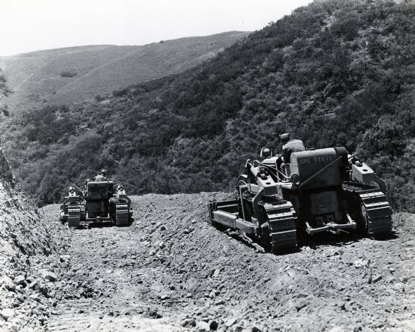 Two men operate International crawler tractors (TracTracTors) with bulldozer blades on a rocky hillside as part of a military training program at Camp Pendleton. The original caption reads: "There is nothing like actual earth moving work on real jobs to develop good bulldozer operators. In Camp Pendleton the need for more and more roads and more camp sites is giving men in training and maintenance groups an experience in crawler-tractor and bulldozing operation that cannot be beat. This series shows a number of Model TD-18 and TD-14 International Diesel crawler-tractors at work on a mountainous road called San Juan Trail which is 14 miles long and connects Camp No. 2 with Casey Springs where a new camp was planned."