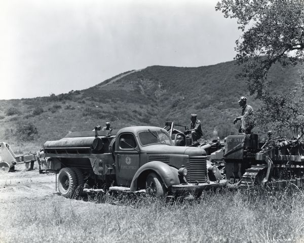 Uniformed men sit on top of International crawler tractors equipped with bulldozer blades in an area of brush with hills in the background. An international truck marked, "Camp Pendleton, USMC 39803" is parked in the foreground.