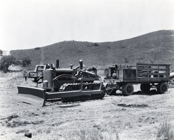 A man uses an International crawler tractor (TracTracTor) to pull a greasing wagon across rough terrain. A dirt road and hills are in the background.
