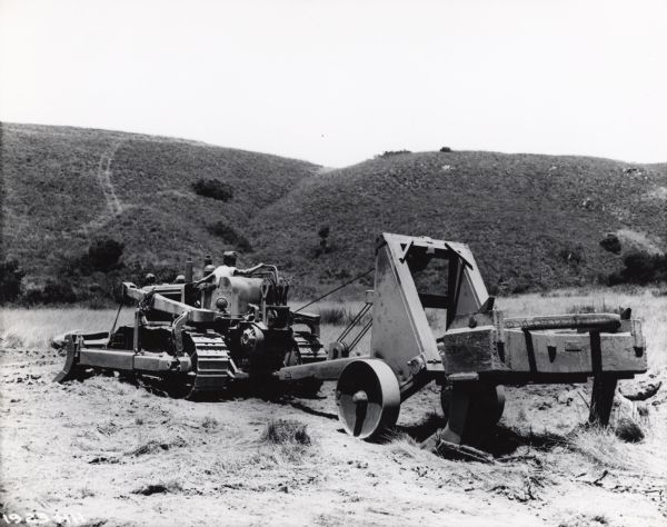 A man uses an International TD-18 crawler tractor (TracTracTor) with a heavy-duty road ripper to do construction on rough terrain. Hills are in the background.